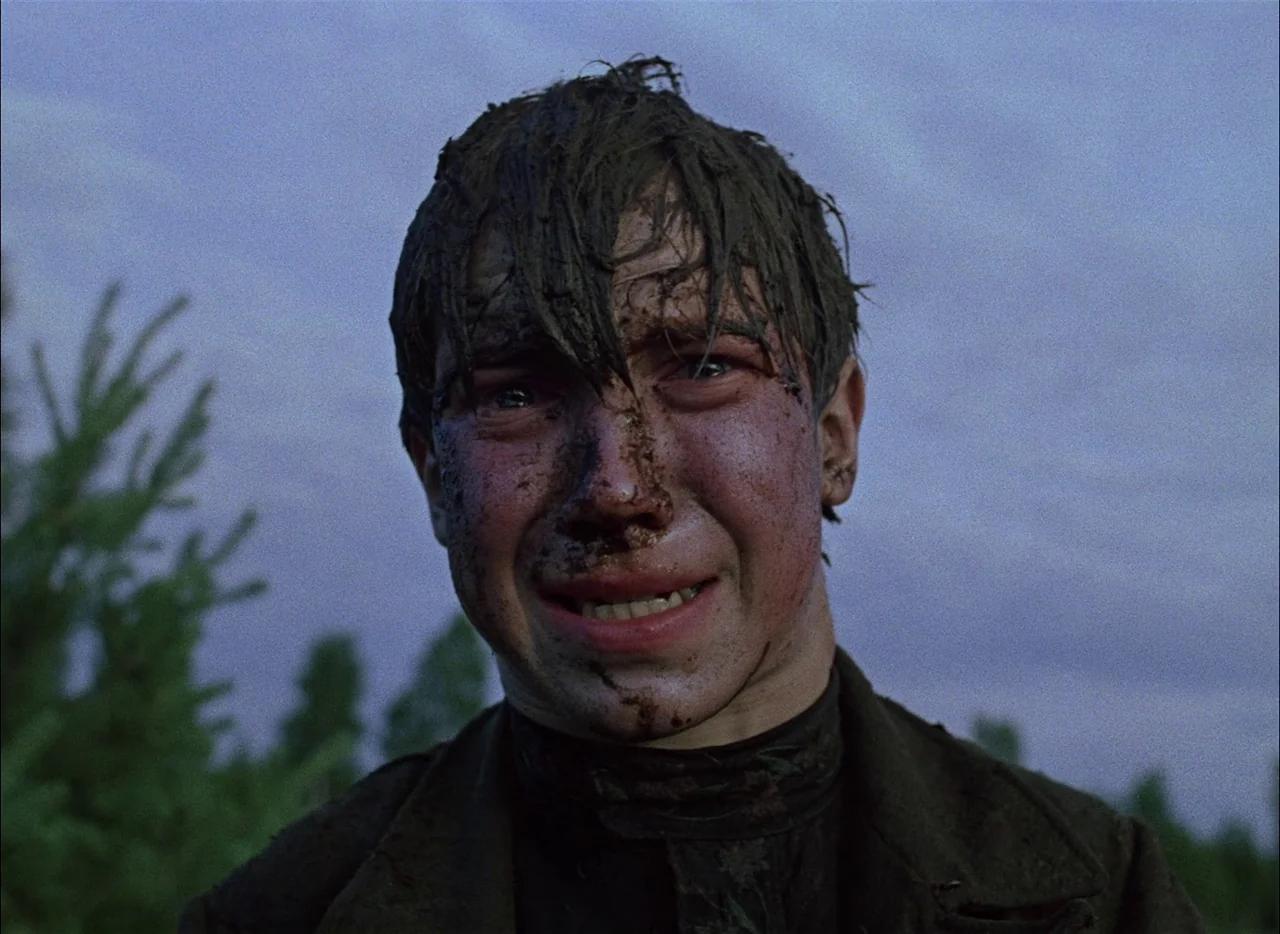 Come and See (1985) movie still: closeup — An open outdoor area at twilight with blurred greenery visible in the distance. A teenager with mud-caked…