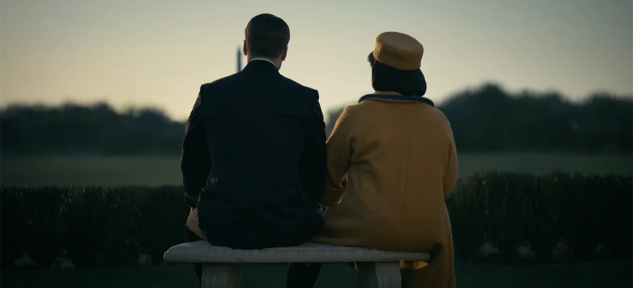 Devotion (2022) movie still: medium — A stone bench sits in an open park with trees and a distant monument silhouette. A man and woman in formal…