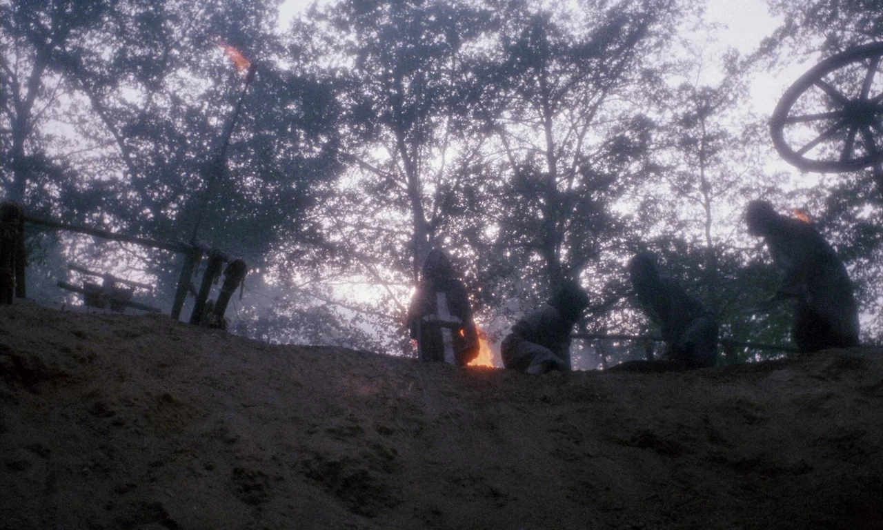 The Church (1989) movie still: wide — A dark dirt ridge at the edge of a silhouetted forest at dusk. Four figures cluster around a small fire as a…