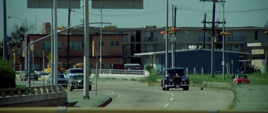 Brewster McCloud (1970) movie still: wide — An urban road stretches past light-colored apartment buildings on a bright, hazy day. Several vintage…