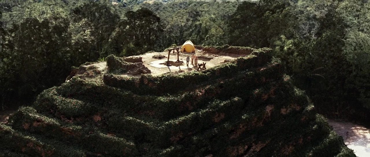 The Ruins (2008) movie still: establishing — A stone stepped pyramid is covered in lush vines and dense jungle foliage. A yellow tent and a group of…
