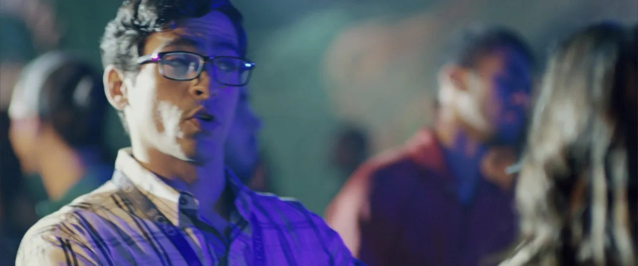 La Musiquita por Dentro (2019) movie still: medium — A crowded, dimly lit indoor space with blurred figures in the background. A young man in…