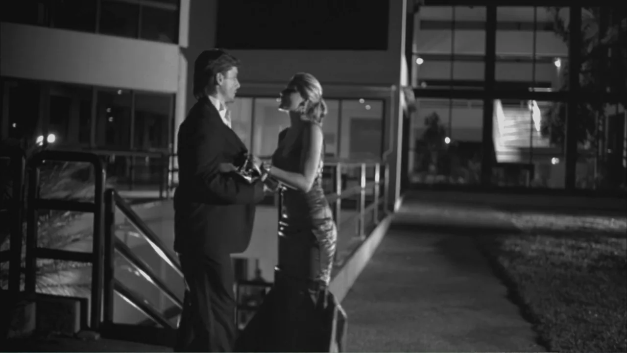 A Distant Place (2020) movie still: fullbody — A modern patio area at night features a glass building wall with interior lighting. A man in a suit…