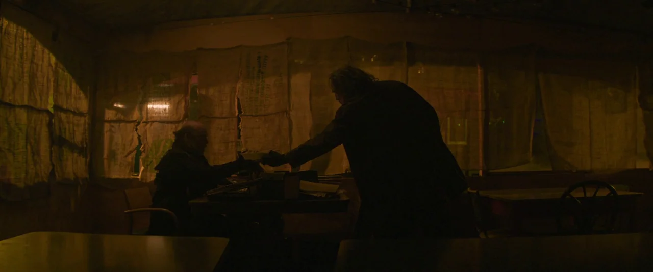 Pig (2021) movie still: medium — A dimly lit room features walls covered with hanging burlap fabric covering the windows. One man stands and hands…