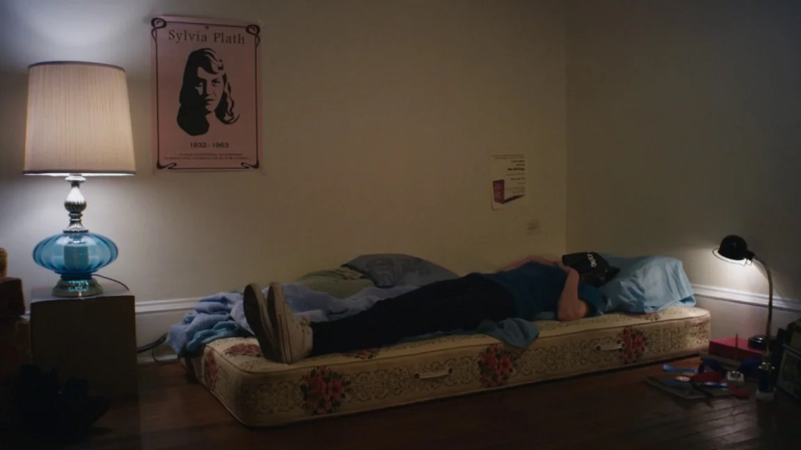 Adult World (2013) movie still: wide — A spare bedroom features a thin mattress on the floor and a Sylvia Plath poster. A person lies motionless on…