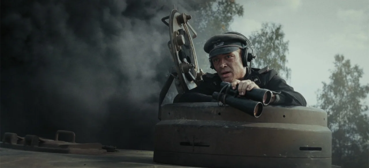 The Tank (2025) movie still: medium — A soldier peers out from the open turret hatch of a military tank. Dark smoke billows in the background behind…