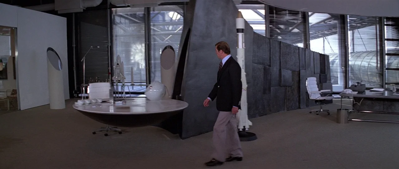 Moonraker (1979) movie still: fullbody — A spacious, minimalist corporate interior features dark geometric wall panels and large glass windows. A…