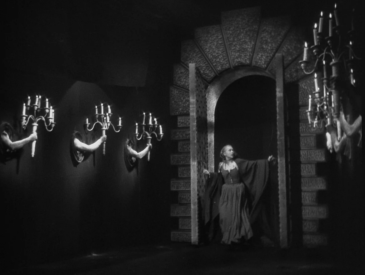 La Belle et la Bête (1946) movie still: wide — A dark stone hallway features a large arched doorway and wall-mounted human-like arms holding…