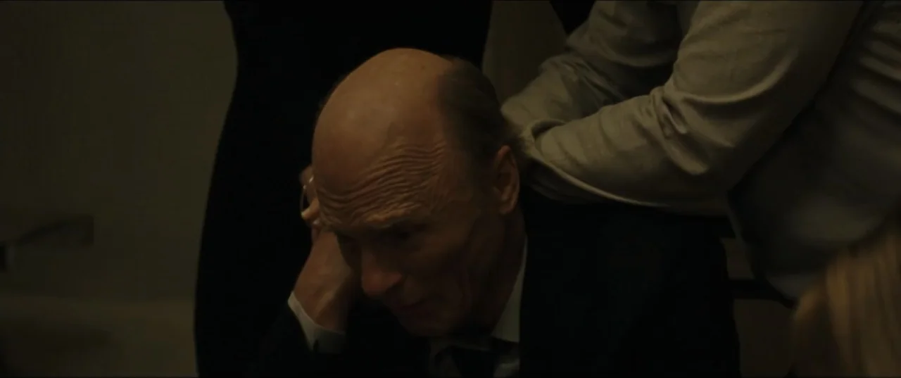 mother! (2017) movie still: closeup — A dimly lit interior space with a person in a suit present. The man stares downward with a hand supporting his…