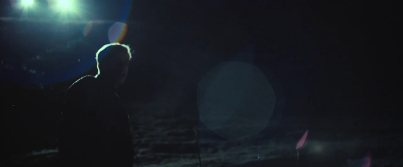 Rift (2017) movie still: medium — An open dark field at night illuminated by harsh lights from off-camera. A lone man in silhouette stands against…