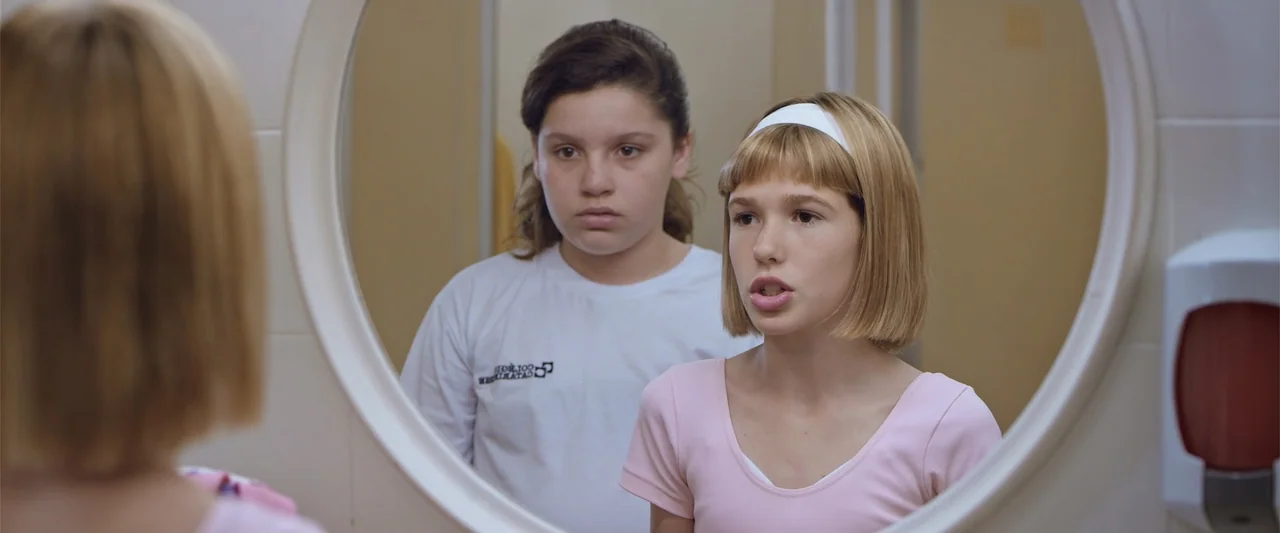 Dirty Little Secret (1998) movie still: medium — Two young girls are positioned in front of a circular bathroom mirror. A girl in a pink shirt…