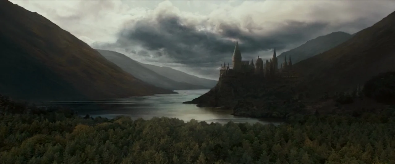 A stone castle complex sits on a high cliff overlooking a wide lake. Massive steep mountains flank the water under a dense, overcast, cloud-filled sky.