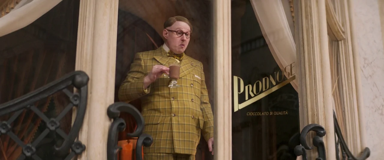 Wonka (2023) movie still: medium — A storefront with glass panes and elegant gold lettering displays the name Prodnose. A man in a patterned yellow…