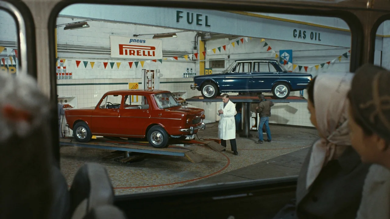 Playtime (1967) movie still: wide — An auto repair shop with white tiled walls features two cars on hydraulic lifts. A mechanic inspects a red car…