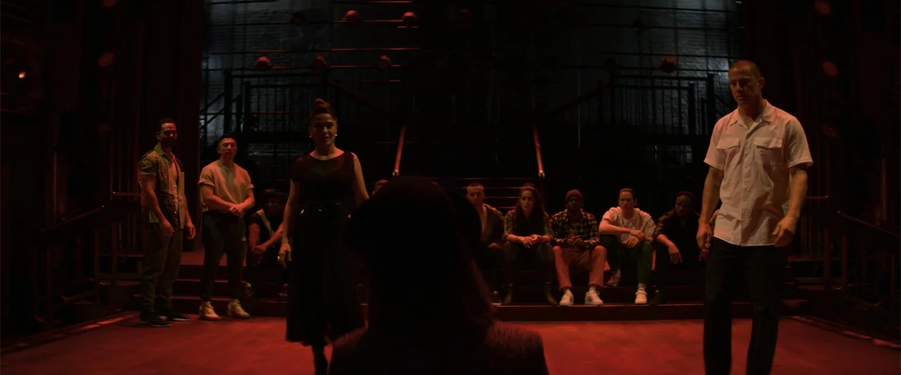 Magic Mike's Last Dance (2023) movie still: wide — A dark wooden stage features a group of people sitting on tiered risers behind. A woman and a man…