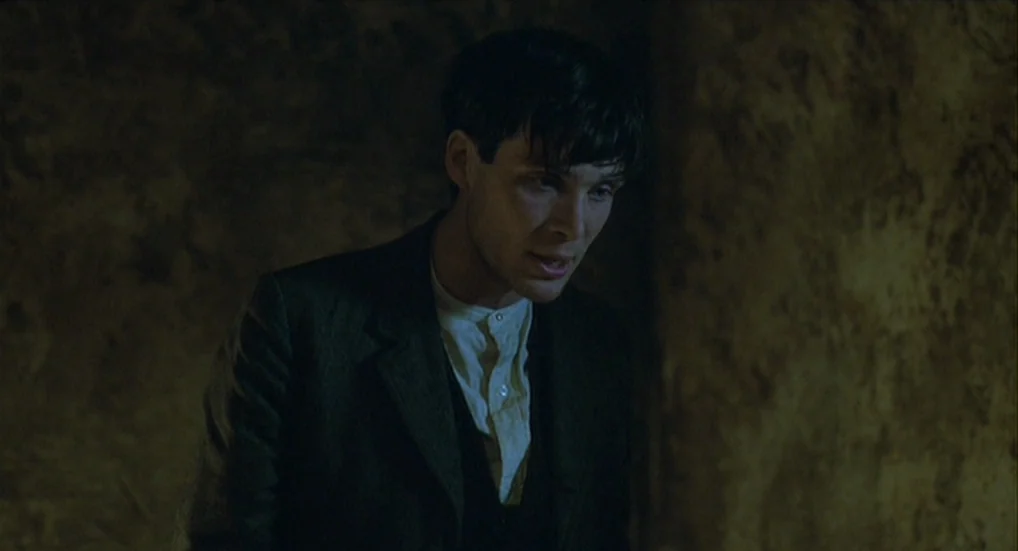 The Wind That Shakes the Barley (2006) movie still: medium — A dark, dimly lit room with textured, uneven walls. A man in a suit looks down while…