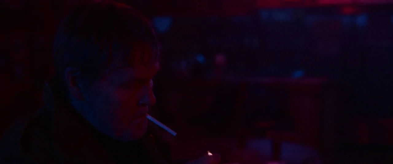 VFW movie still: closeup — A dimly lit space is dominated by intense red and blue light. A man holds a cigarette to his lips while lighting it with…