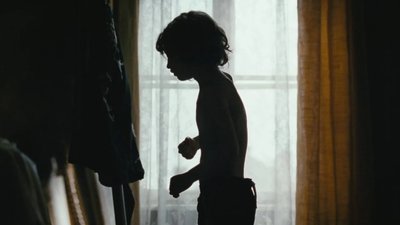Childhood of a Leader (2015) movie still: medium — A room with lace window curtains and a standing clothing rack in the foreground. A shirtless boy…