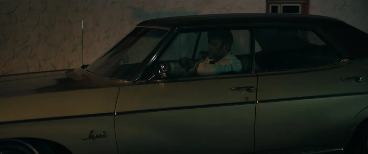 Guns Up (2025) movie still: medium — A dark, gold-colored vintage sedan parked next to a textured stucco wall at night. A man sits in the driver…
