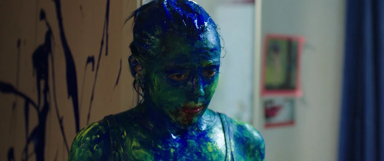 Raw (2016) movie still: medium — A dimly lit room contains walls with dark paint splatters and blurred photographs. A young woman covered in thick…