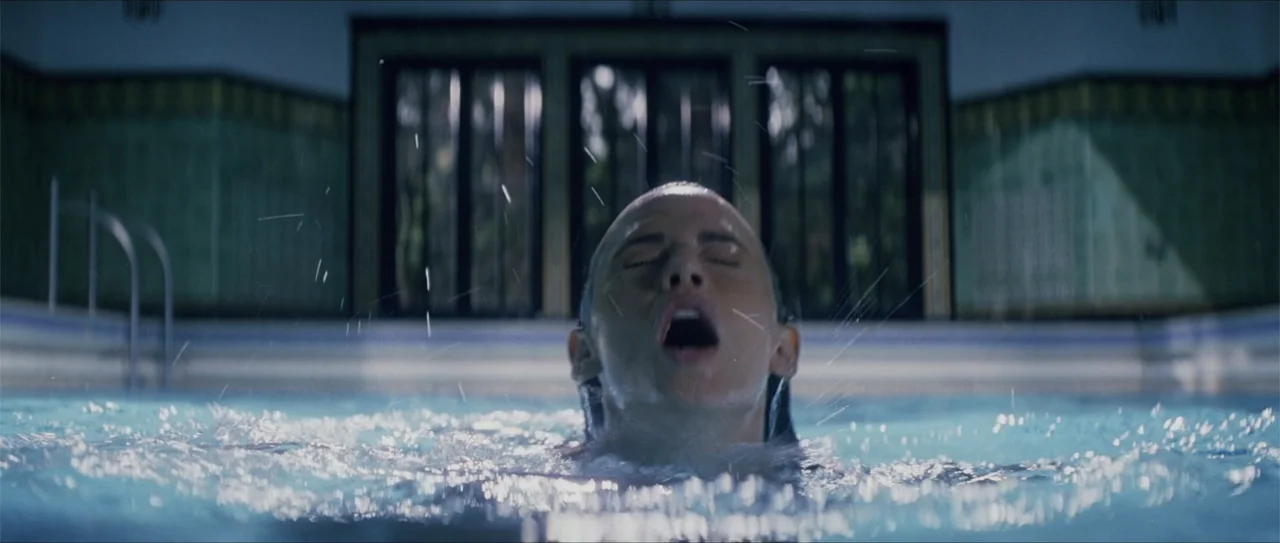 Elektra (2005) movie still: closeup — A dimly lit indoor swimming pool features tiled walls and dark windows in back. A young woman emerges from the…