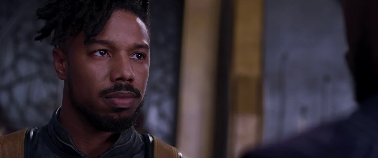 Black Panther (2018) movie still: closeup — A dimly lit indoor space with textured gray walls and vertical architectural pillars. A man with styled…