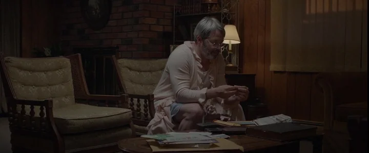 To Dust - Nightmares (2018) movie still: medium — A 1970s interior with dark wood-paneled walls and a brick fireplace. A middle-aged man in a pink…