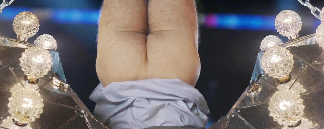 The Naked Gun (2025) movie still: closeup — A person performs an inverted handstand above two mirrored, lit prop structures. Blurred blue and…