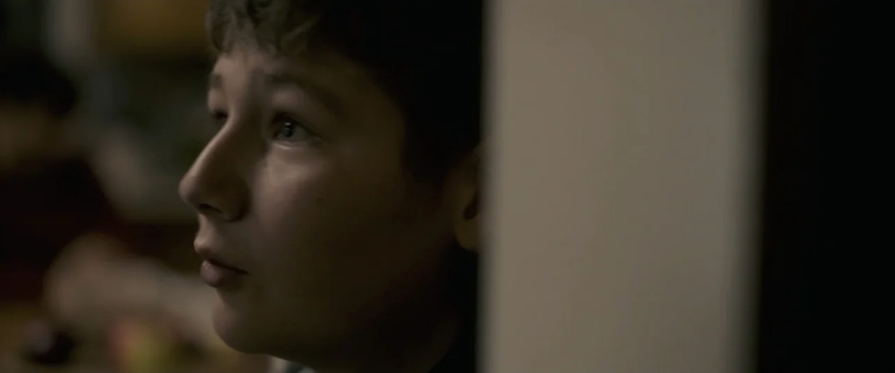 Talk to Me (2023) movie still: closeup — A dimly lit indoor space with a vertical wall partition dividing the frame. A young boy stands behind the…