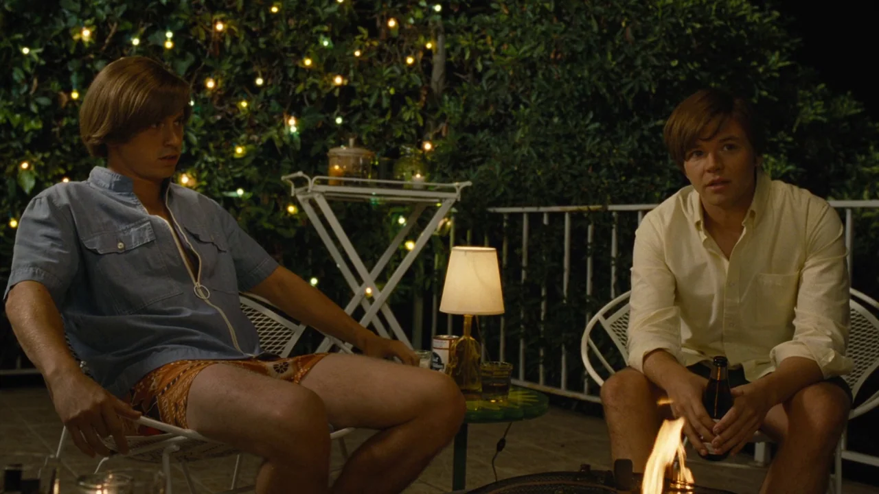 Love & Mercy (2014) movie still: medium — A night patio space features two men sitting before a lit fire pit. Behind them is a hedge illuminated by…