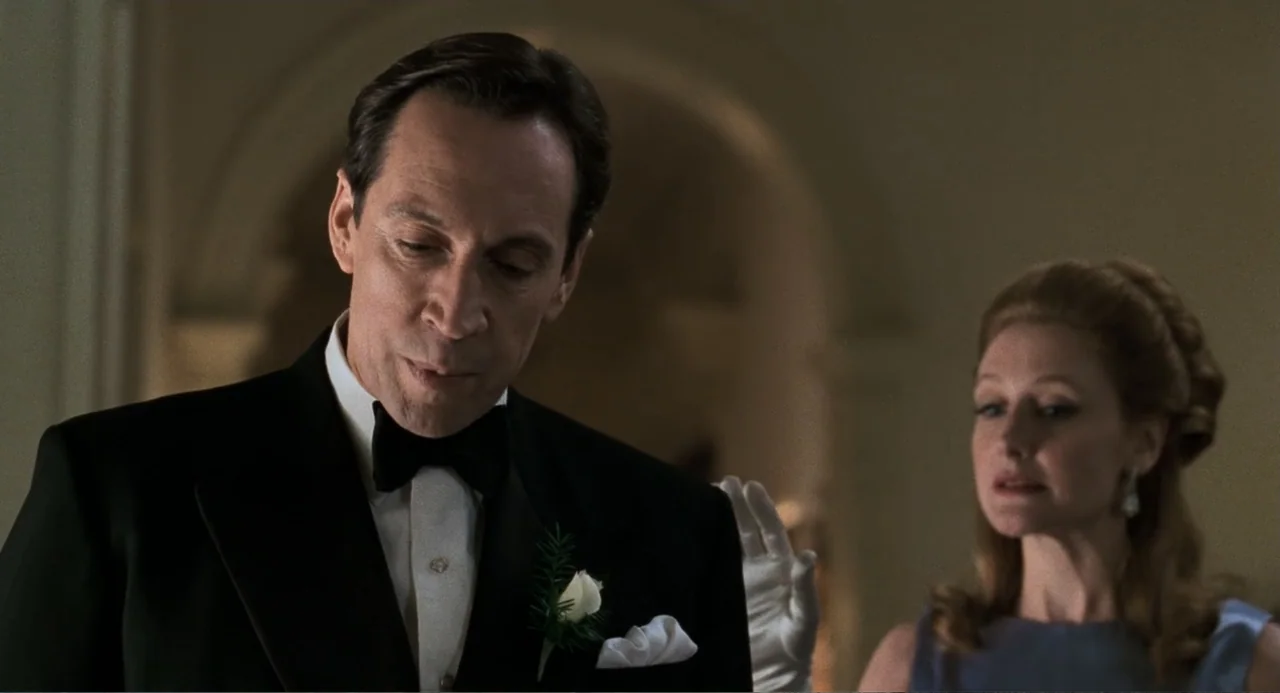 Jumanji (1995) movie still: medium — A well-lit formal interior with light-colored walls and arched architectural features. A man in a tuxedo stands…