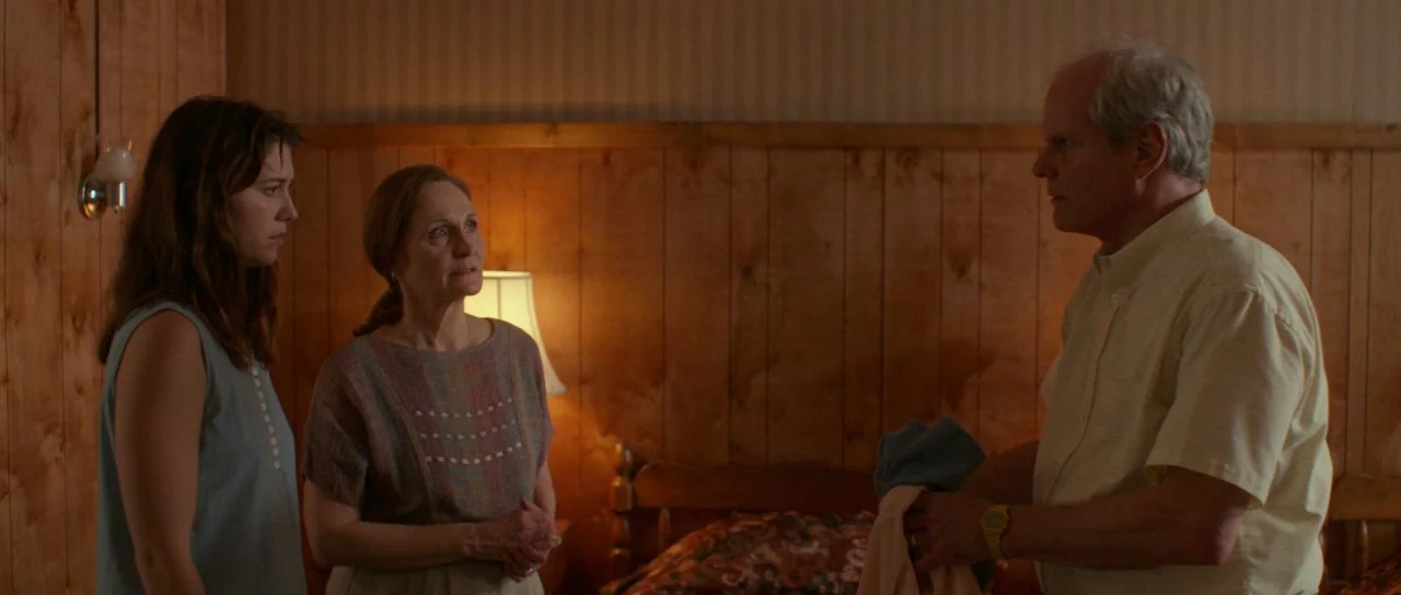 Faults (2014) movie still: medium — A dimly lit room features wood-paneled walls and a lamp on a bedside stand. Two women and one man stand near a…