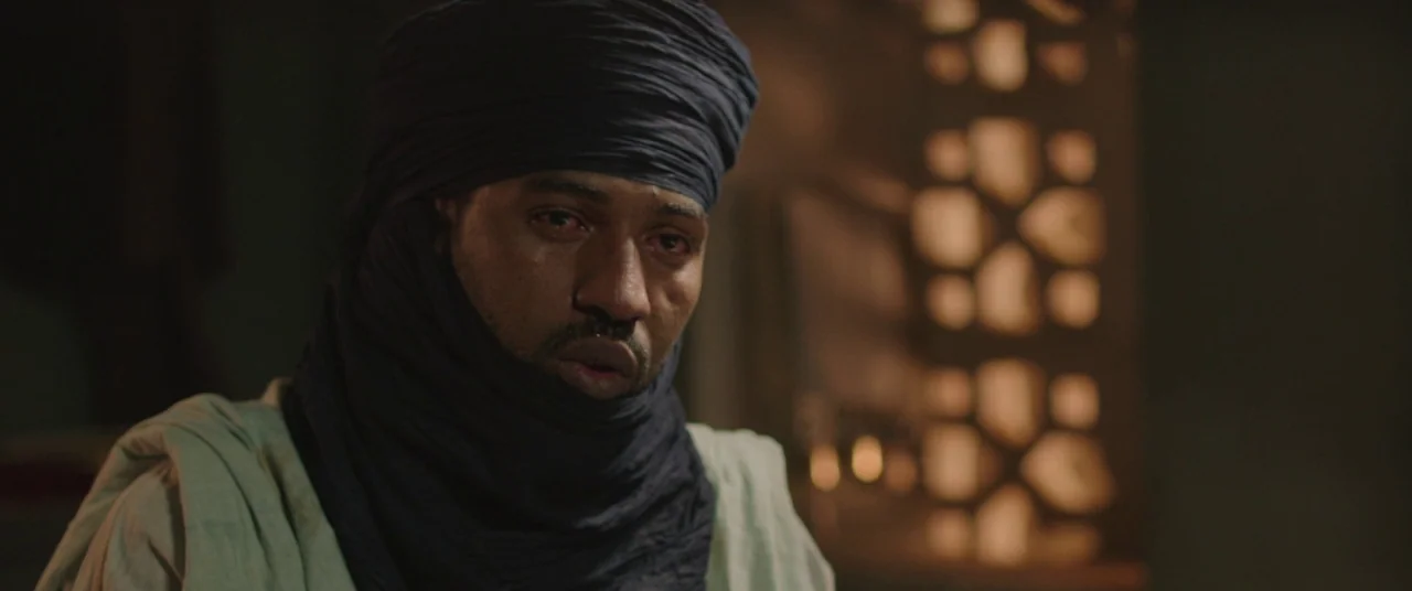 Timbuktu (2014) movie still: closeup — A dimly lit room with a patterned screen structure visible in the background. A man wears a dark head wrap…