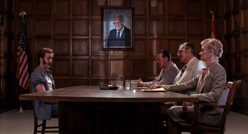 Raising Arizona (1987) movie still: wide — A boardroom with dark wood-paneled walls and a large, central wooden meeting table. Four people sit at…