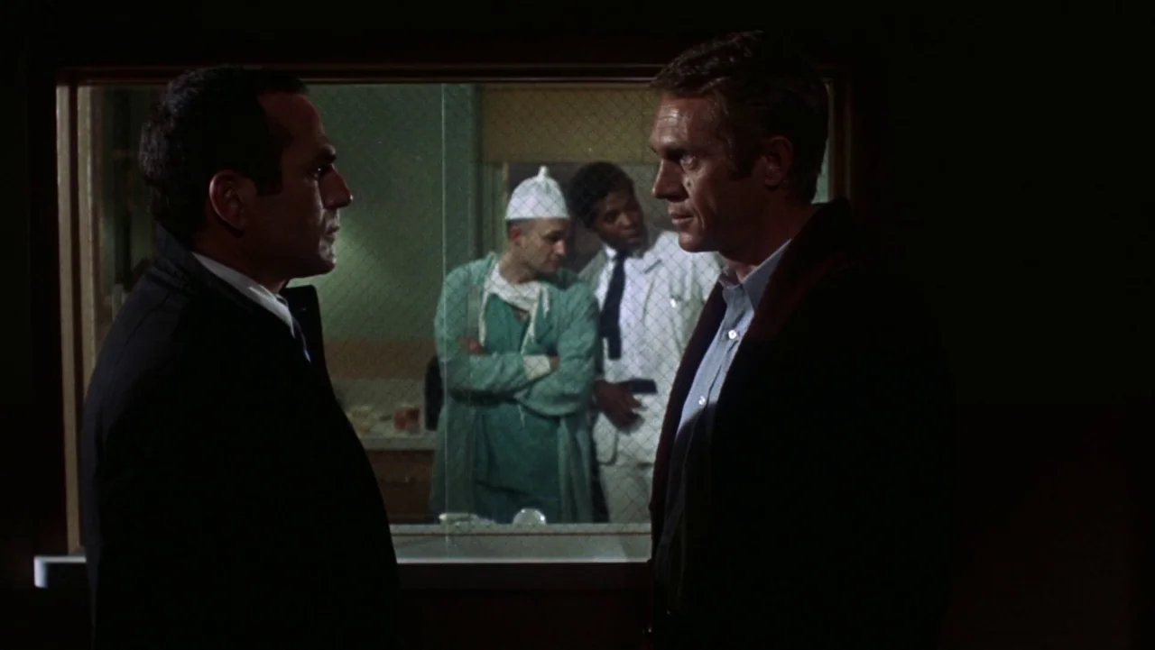 Bullitt (1968) movie still: medium — A dimly lit hospital corridor with a glass window looking into a sterile area. Two men in dark suits converse…