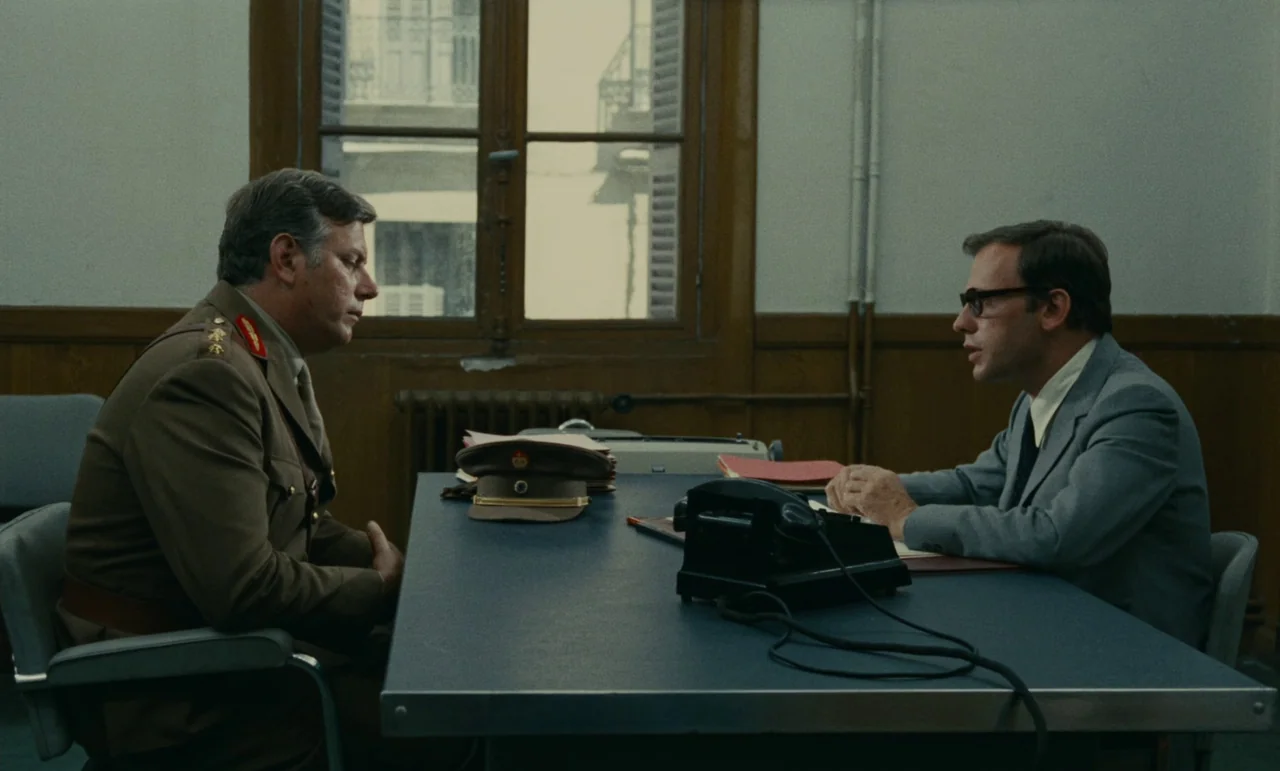 Z (1969) movie still: medium — A sparse, functional office features wood paneling and a large window with shutters. A military officer and a…
