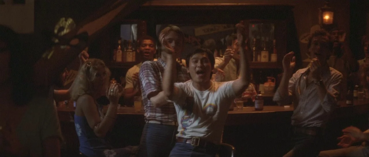 Take Me Away! (1978) movie still: medium — A dimly lit tavern with a dark wooden bar and shelves of bottles. Several patrons in casual 1980s attire…