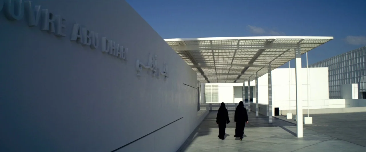 6 Underground (2019) movie still: wide — A modern white building complex with an overhead geometric canopy structure in Abu Dhabi. Two women in…