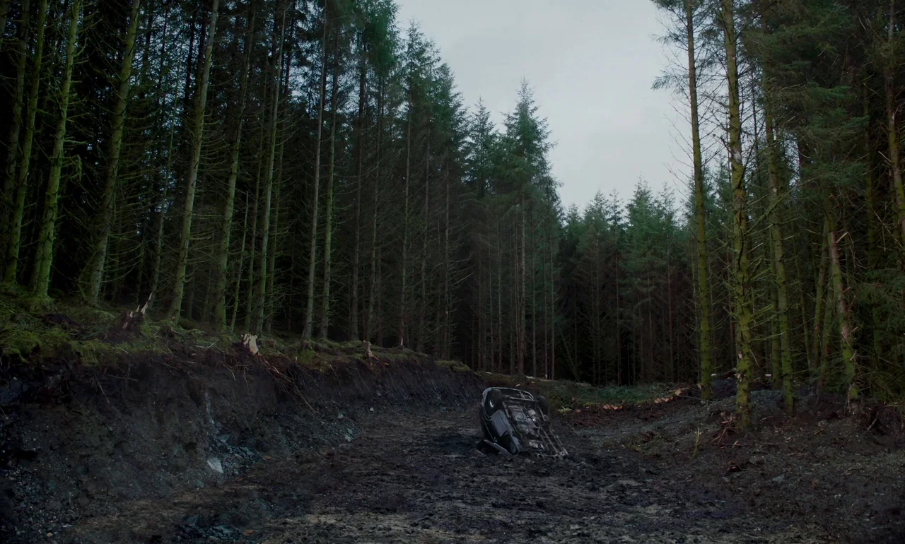 It Is In Us All (2022) movie still: wide — A dense, overgrown pine forest stands beside a deep, muddy dirt excavation clearing. A single car lies…