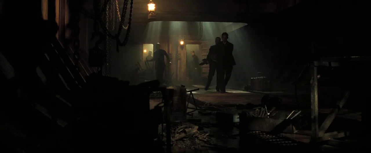 Casino Royale (2006) movie still: wide — A dark cargo hold is filled with hanging chains and scattered metallic equipment. Three men in suits walk…