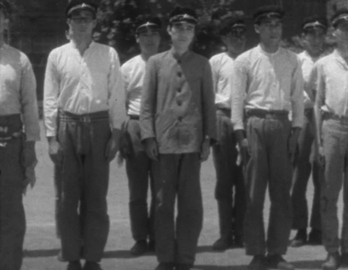Tokyo Chorus (1931) movie still: fullbody — A group of young men stand in a line on a flat, open outdoor area. They are dressed in uniform tunics…
