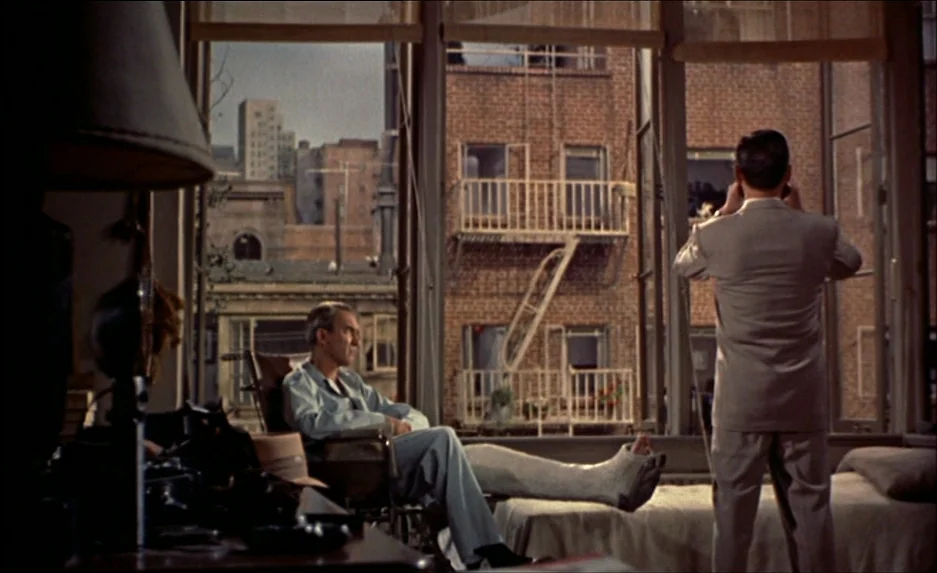 Rear Window (1954) movie still: wide — A spacious apartment room overlooks a red brick building with fire escapes outside. One man sits in a…