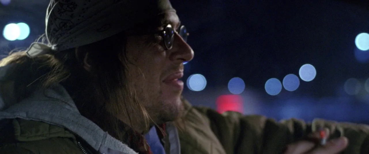 The End of the Tour (2015) movie still: closeup — The interior of a vehicle at night with blurred city lights visible through glass. A man with long…