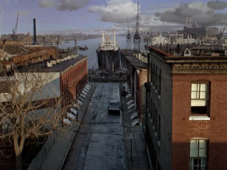 Marnie (1964) movie still: establishing — A city street flanked by brick buildings leads toward a harbor with docked ships. Cranes and industrial…