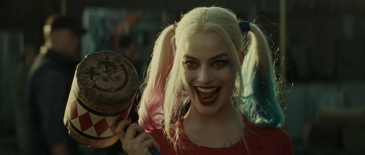 Suicide Squad (2016) movie still: closeup — A character with dip-dyed blonde pigtails stands in an indistinct outdoor setting. She holds a large…