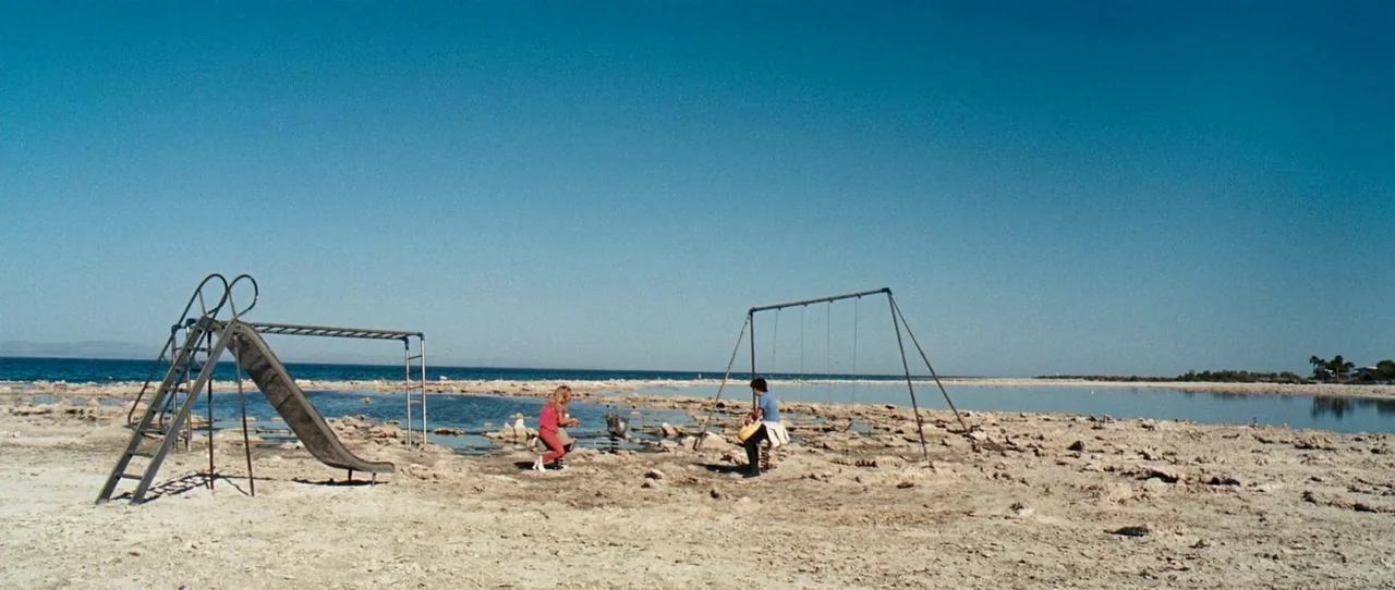 Little Birds (2011) movie still: wide — A desolate shoreline with rocky ground and a playground structure by the water. Two individuals sit on…