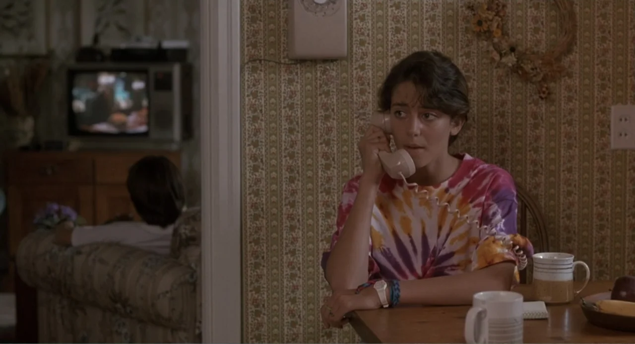 F/X2 (1991) movie still: medium — A residential room features floral wallpaper and a small wooden table in the foreground. A teenage girl sits at…