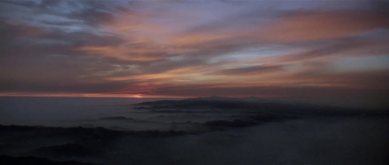 Lost Highway (1997) movie still: establishing — A wide mountain range sits beneath a expansive sky layered with dark, thin clouds. Low-hanging mist…