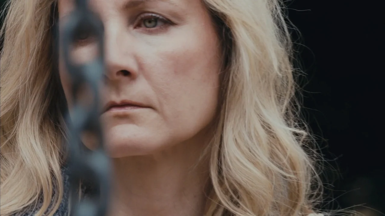 You Might Get Lost (2021) movie still: closeup — A close-up view of a woman's face partially hidden behind a dark metal chain. She gazes ahead with…