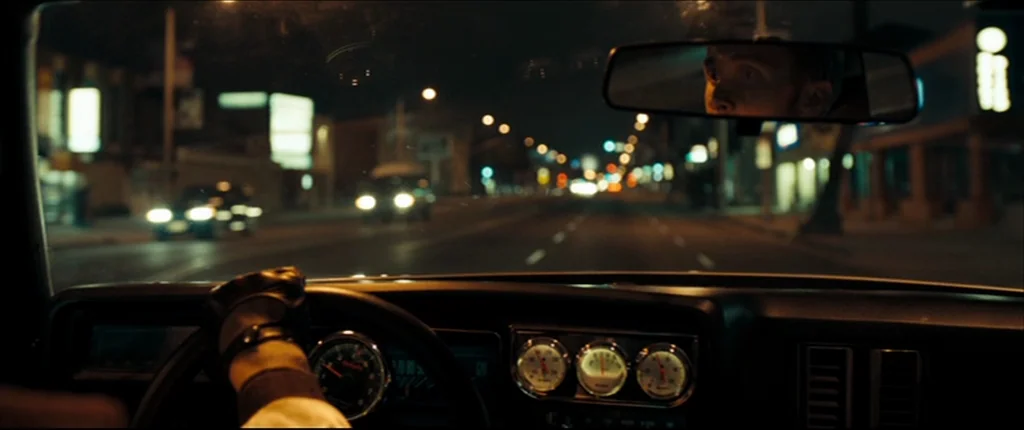 Drive (2011) movie still: medium — A dark, modern car interior at night with glowing analog dashboard gauges visible. The driver is reflected in the…